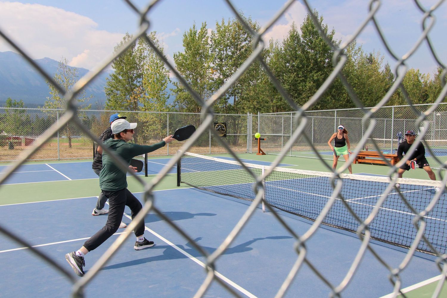 Players enjoy a pickleball match on outdoor blue courts with mountain views through a chain-link fence.