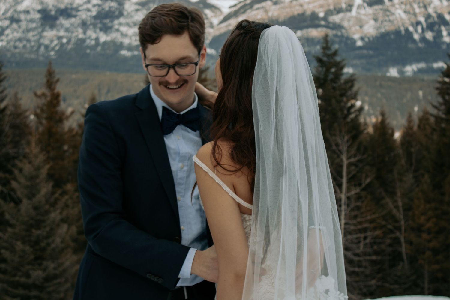 A groom in a navy suit and bow tie smiles at his bride in a white veil against a mountain backdrop.