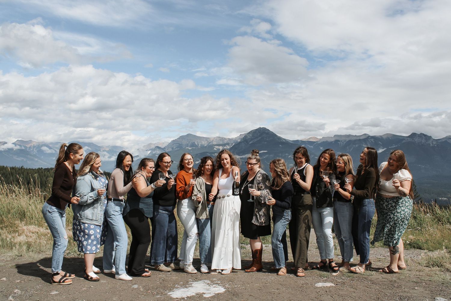 Group of women posing and laughing together on a mountain overlook with scenic peaks and clouds behind them.