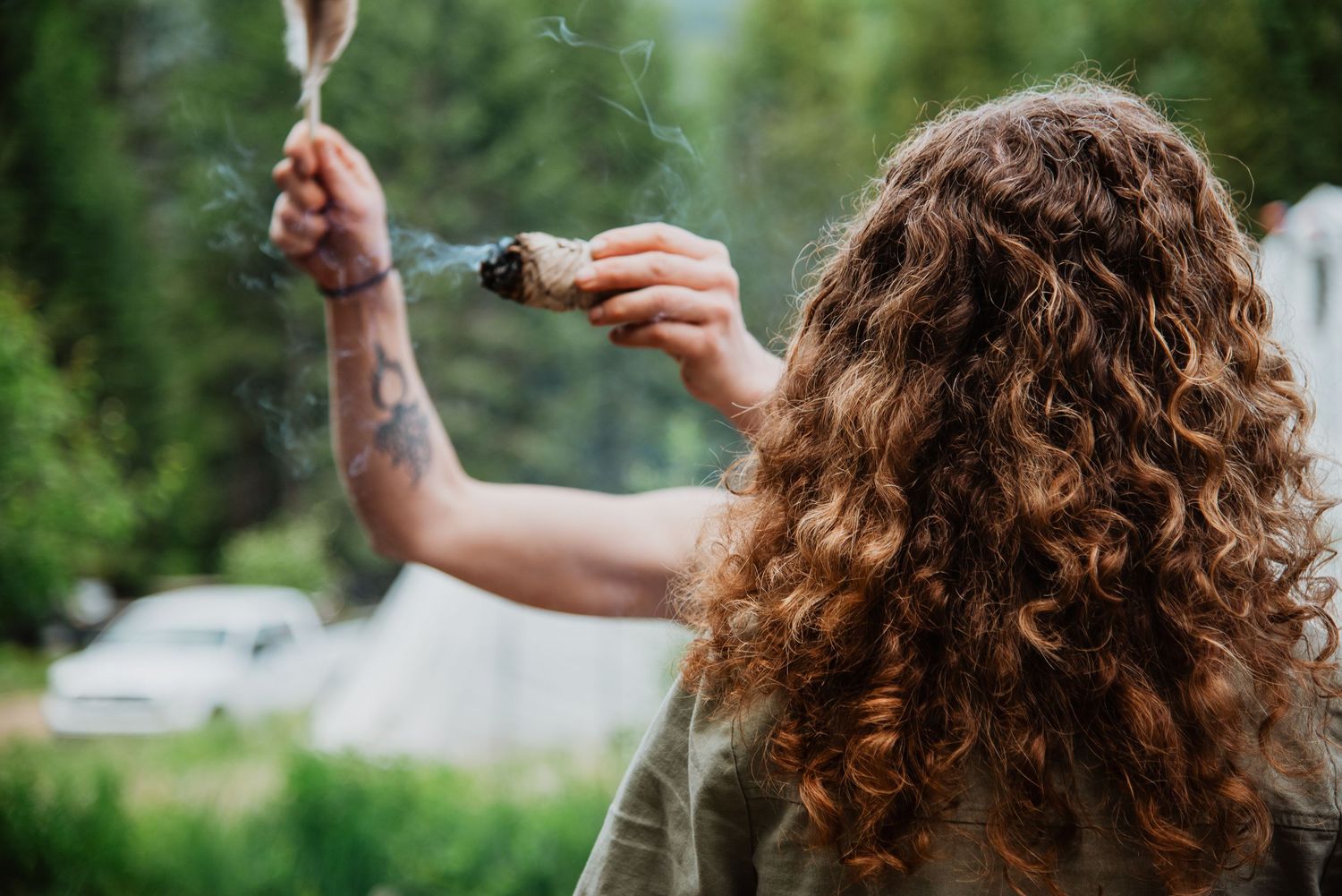Woman with curly hair and tattoo holding burning sage smudge stick outdoors in nature.