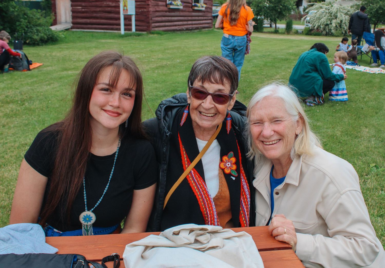 Three women smiling together at an outdoor picnic table on a sunny day at a community gathering.