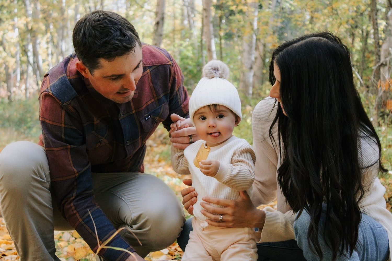 A young family enjoys an autumn outdoor photo session with their baby in a white knit hat among golden fall foliage.
