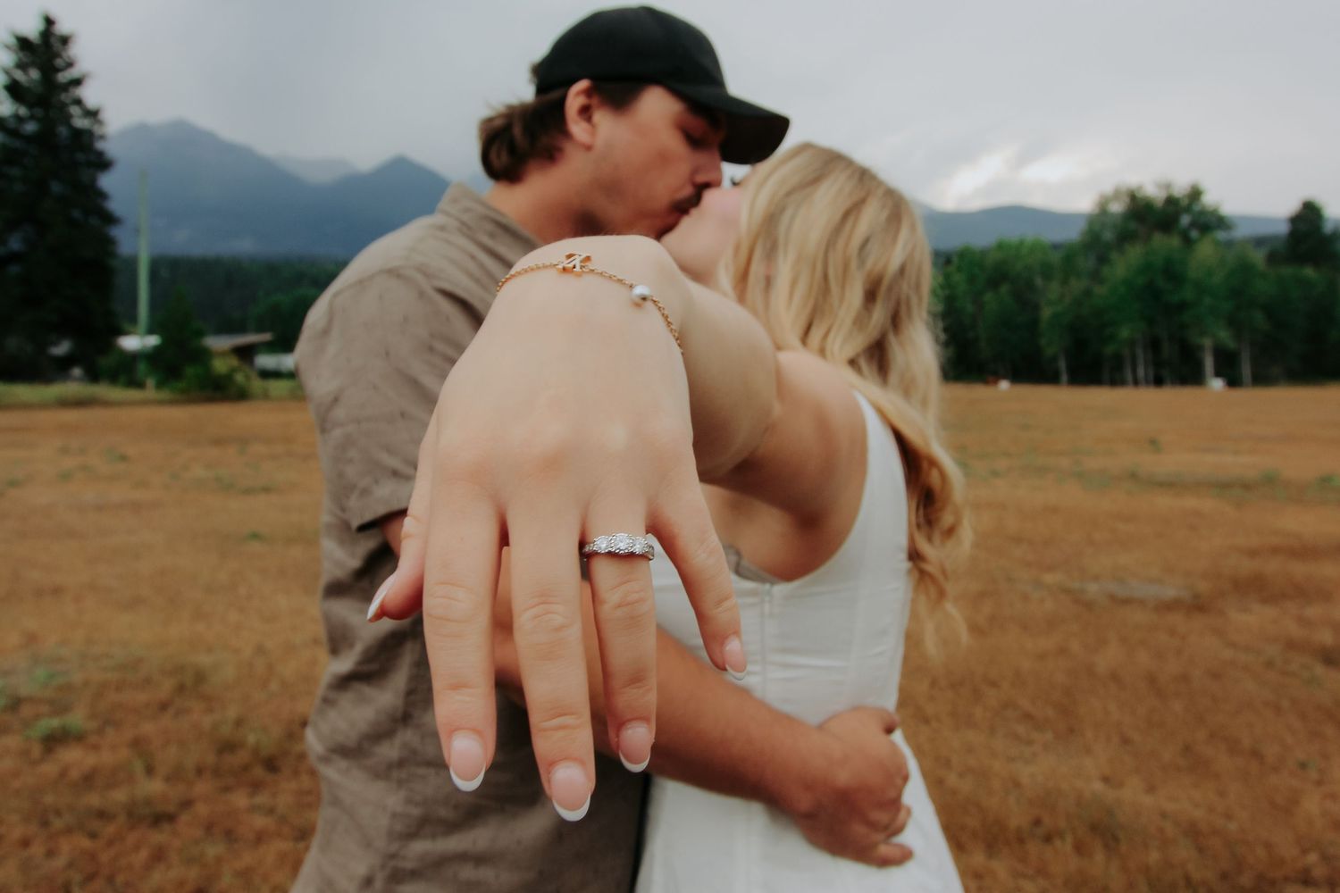 Couple embracing in a field with mountains, woman showing off engagement ring on her hand.