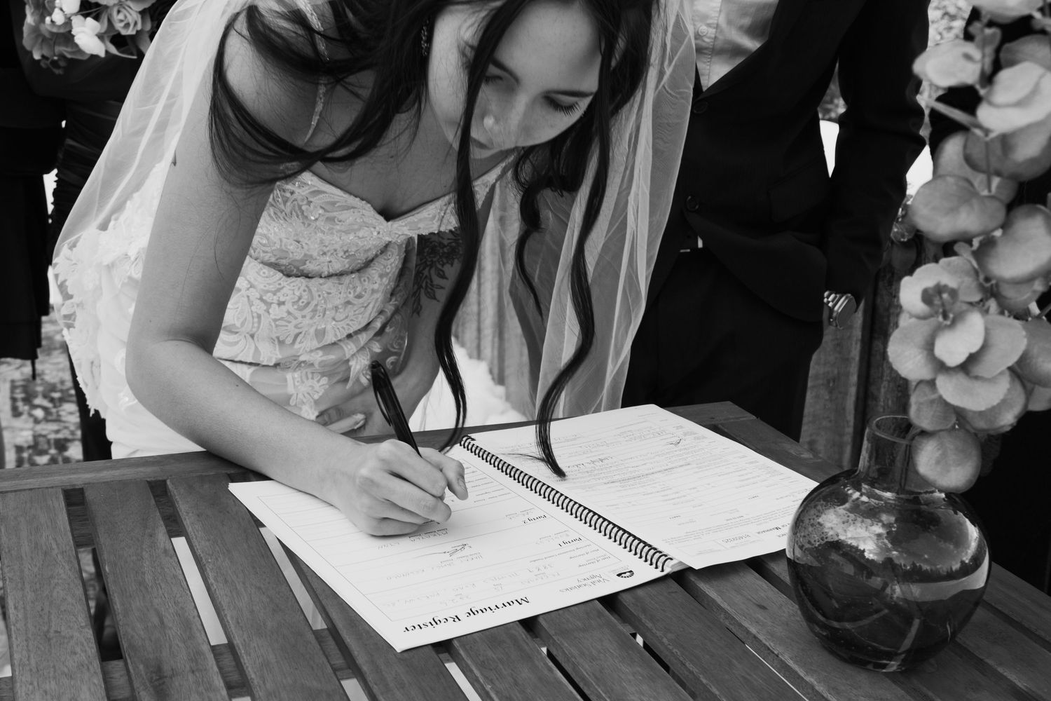 Bride in lace wedding dress signing marriage register at outdoor ceremony table in black and white photo.