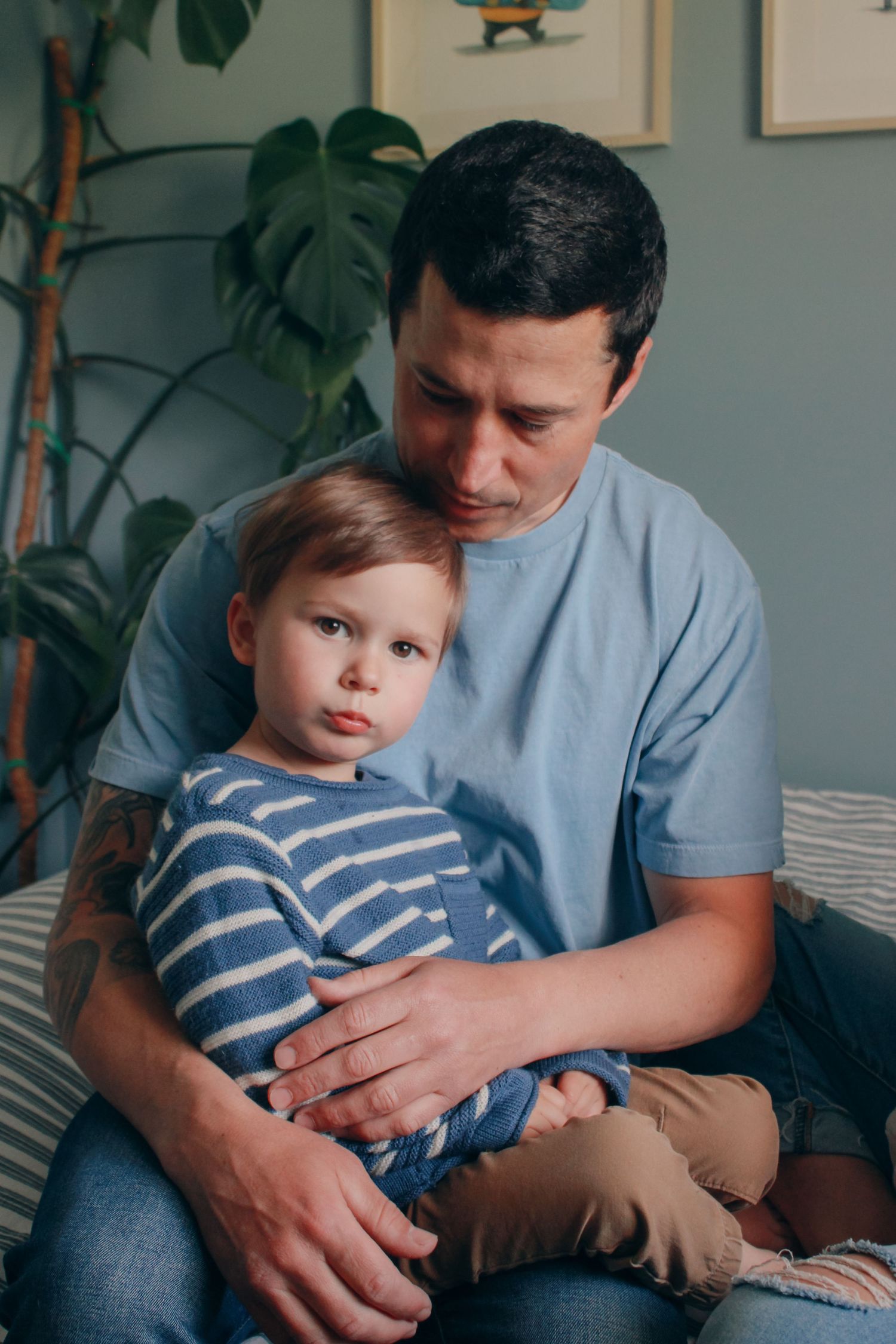 A father tenderly embraces his young toddler son while sitting together on a couch at home.