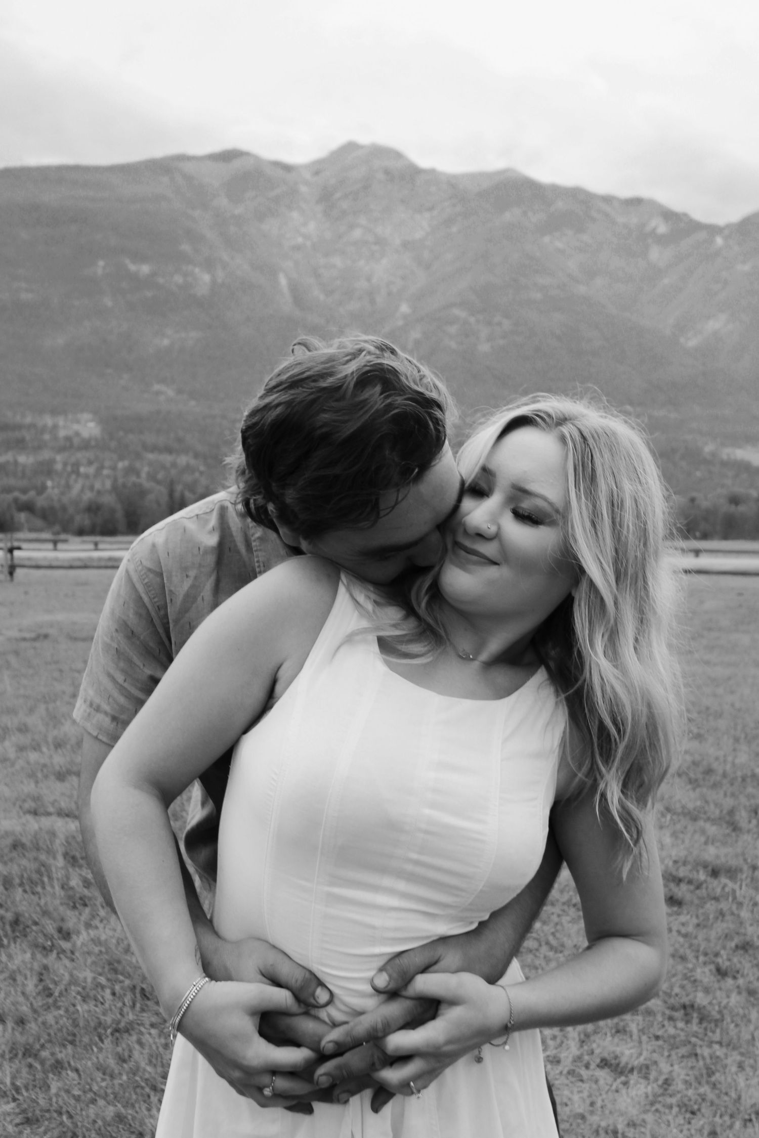 Black and white photo of a couple embracing in a mountain meadow, man kissing womans cheek as she smiles softly.