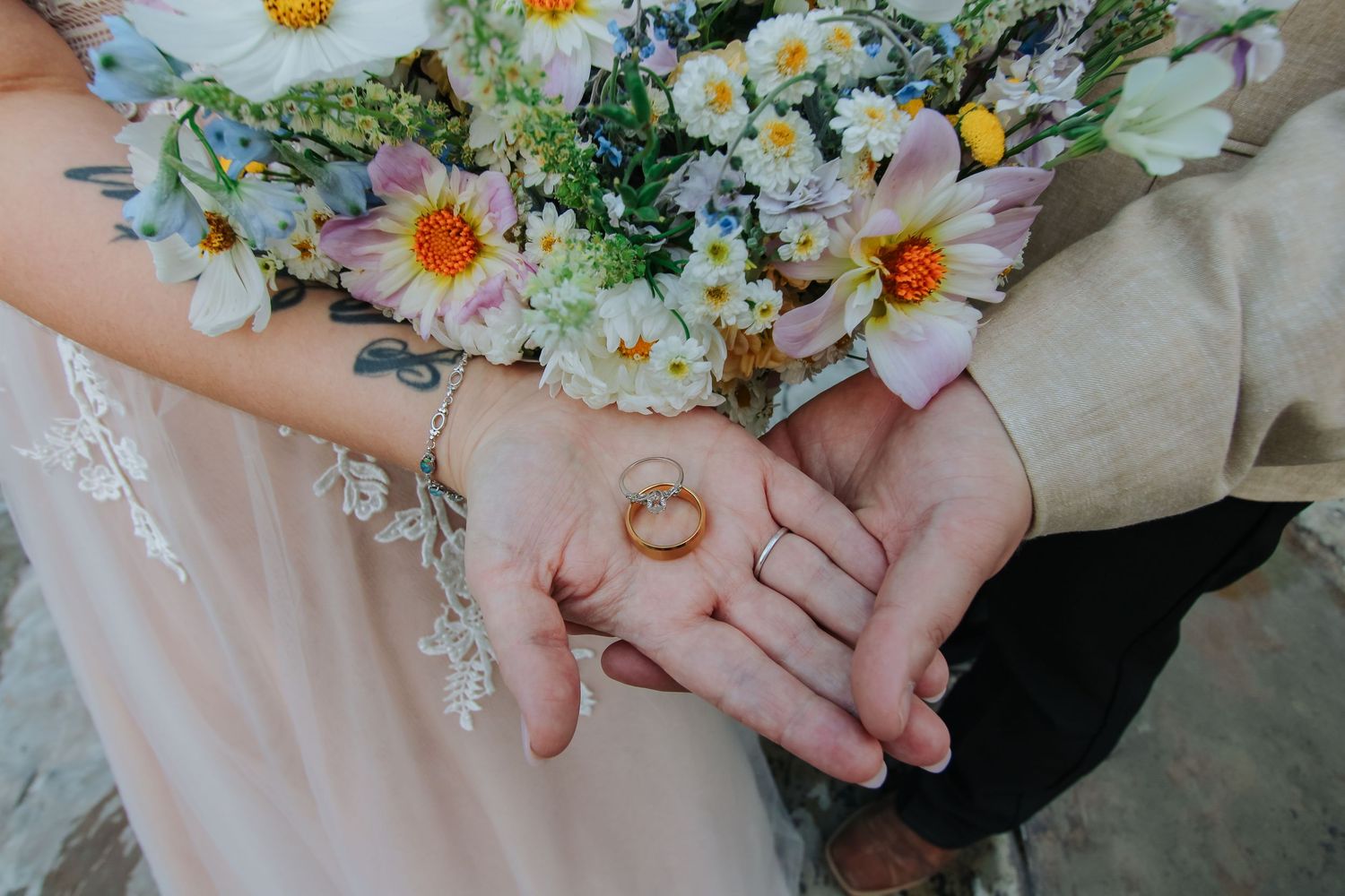 Bride and groom holding gold wedding rings with a colorful wildflower bouquet in the background.