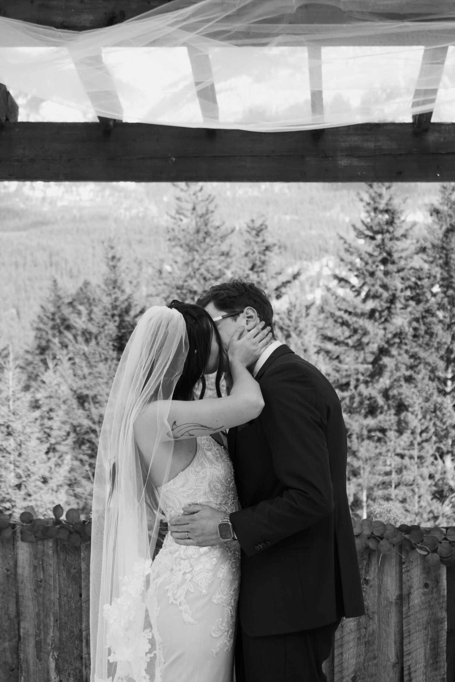 Black and white photo of bride and groom sharing first kiss under pergola with mountain forest backdrop.