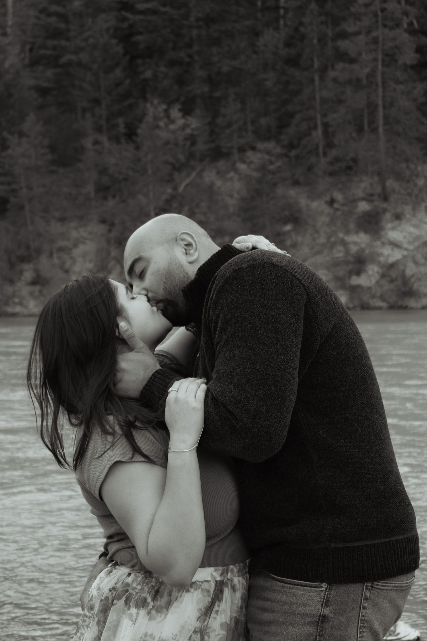 Black and white photo of a couple sharing a romantic kiss near a river with evergreen trees in the background.