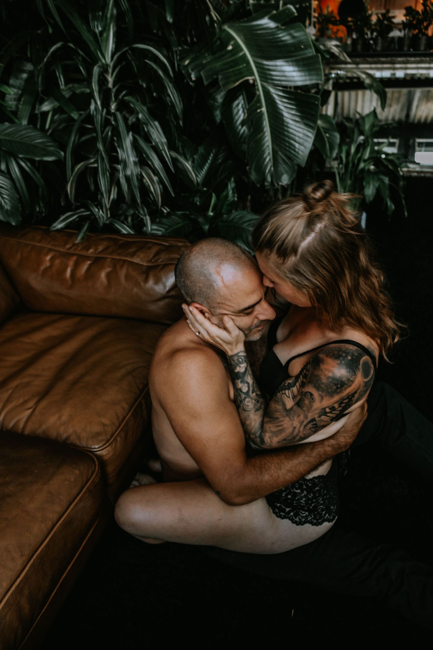 Couple in intimate embrace on leather couch with greenery and dim lighting creating atmospheric mood.