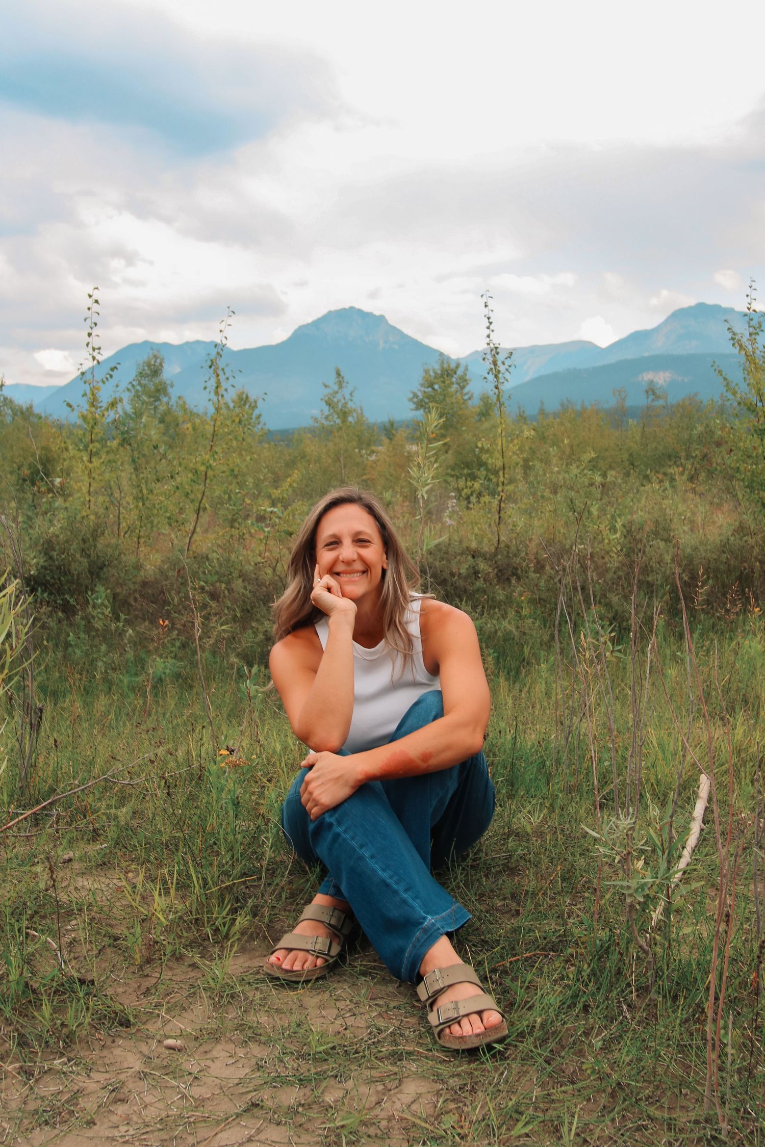 A smiling woman sits cross-legged on grass in jeans and white tank top, with mountains and green trees in the background.