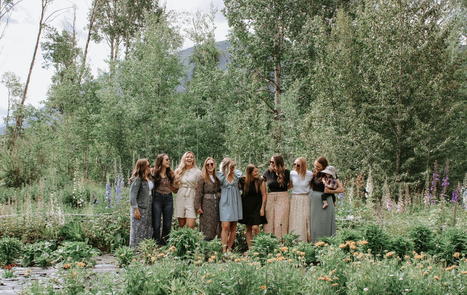 Group of women posing together in a lush garden surrounded by wildflowers and tall trees in a scenic outdoor setting.
