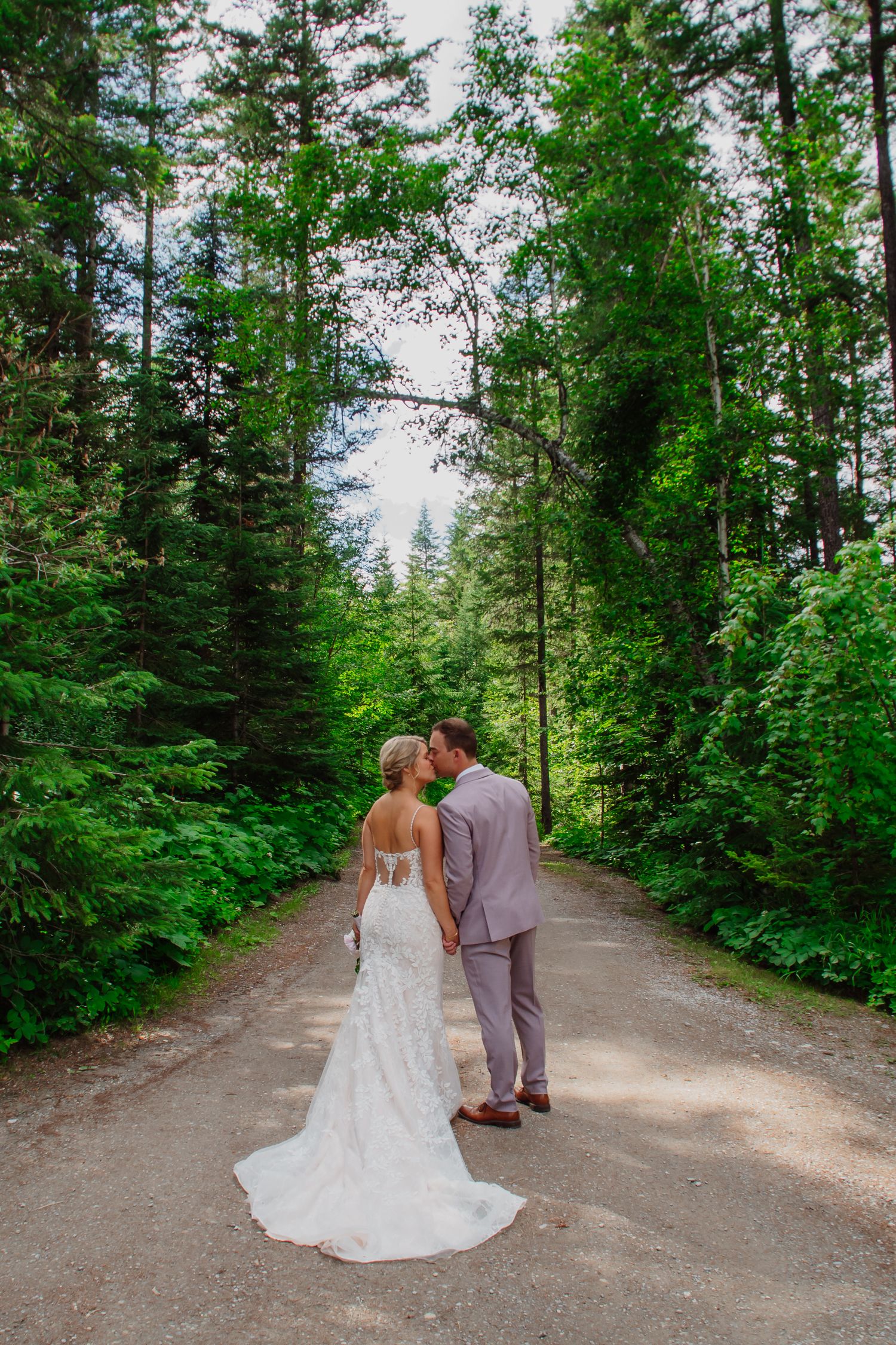 Bride and groom sharing a kiss on a forest path, surrounded by tall green trees, bride in white gown with long train.