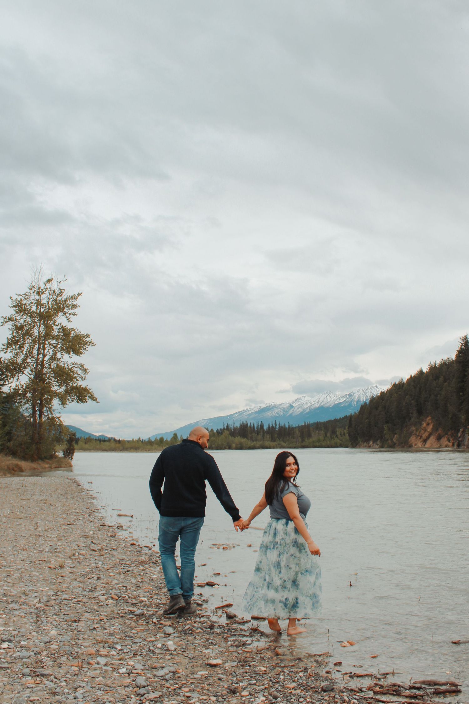 Couple holding hands walking along a rocky riverbank with mountains and evergreen trees in the background.