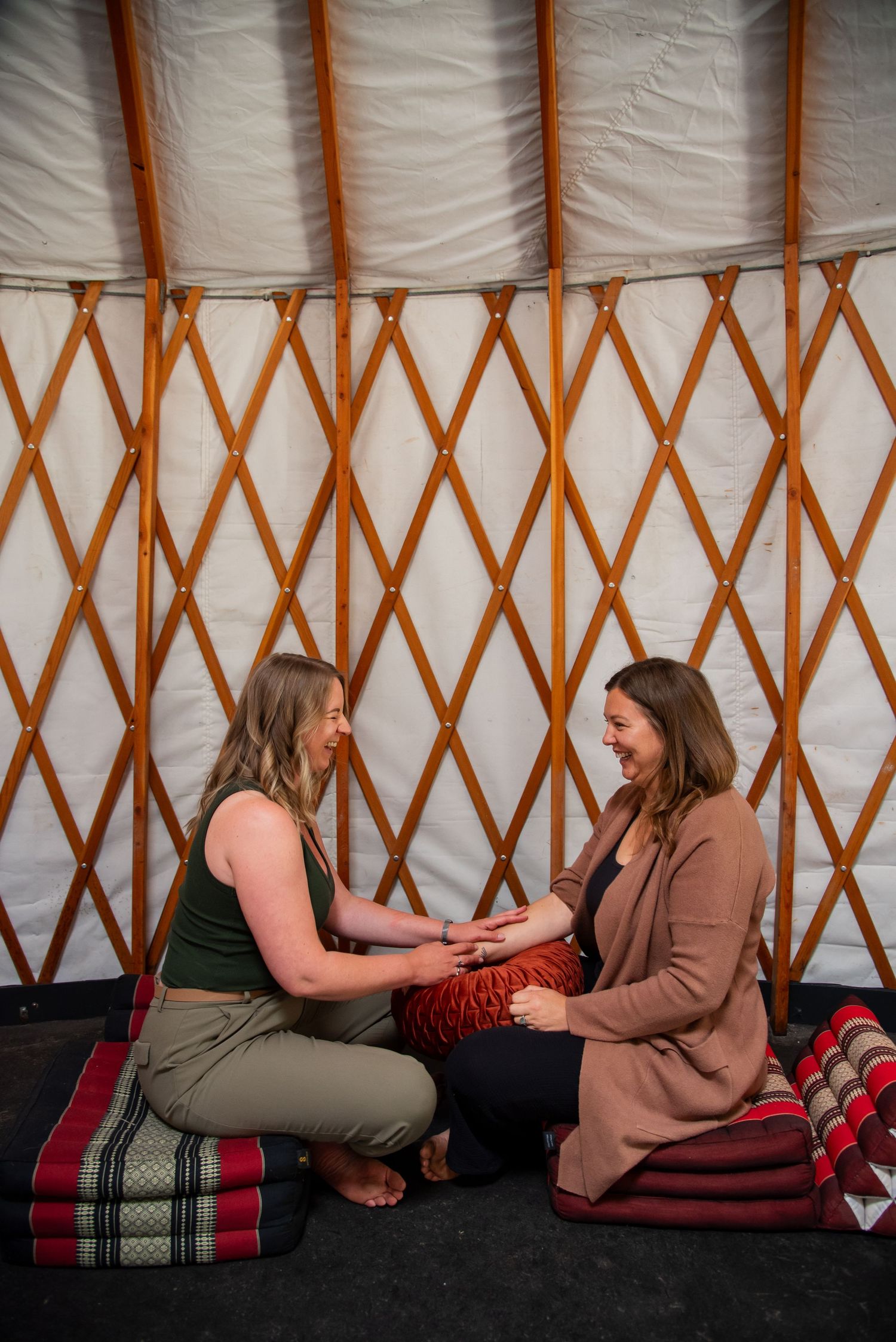 Two women sitting on floor cushions inside a yurt, smiling and holding hands in a warm, intimate setting.