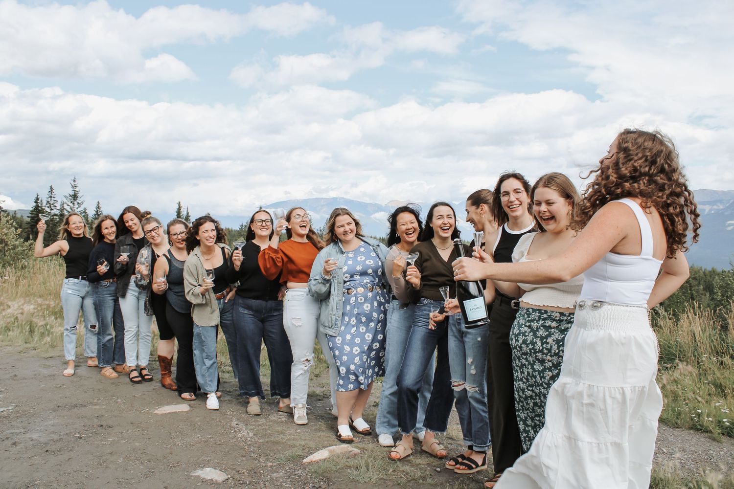 Group of women laughing and celebrating outdoors with wine bottles against a scenic mountain backdrop.