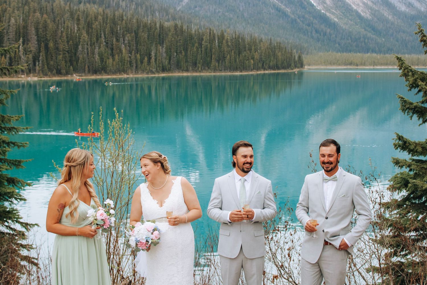 Wedding party in white and light gray attire poses against a scenic mountain lake backdrop.