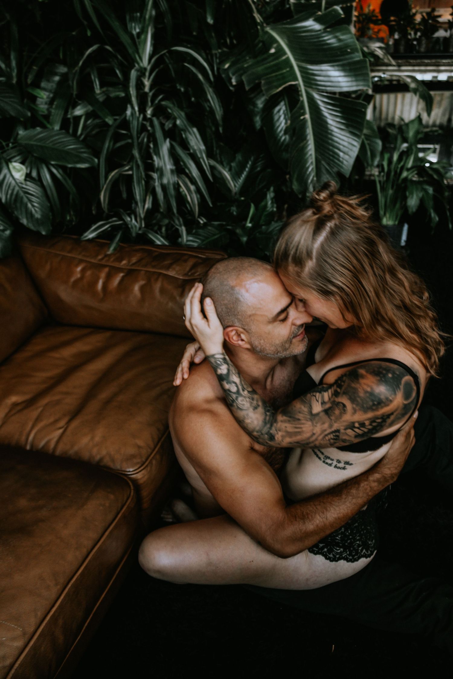 Intimate couple sharing tender moments on a brown leather couch with tropical plants in the background.