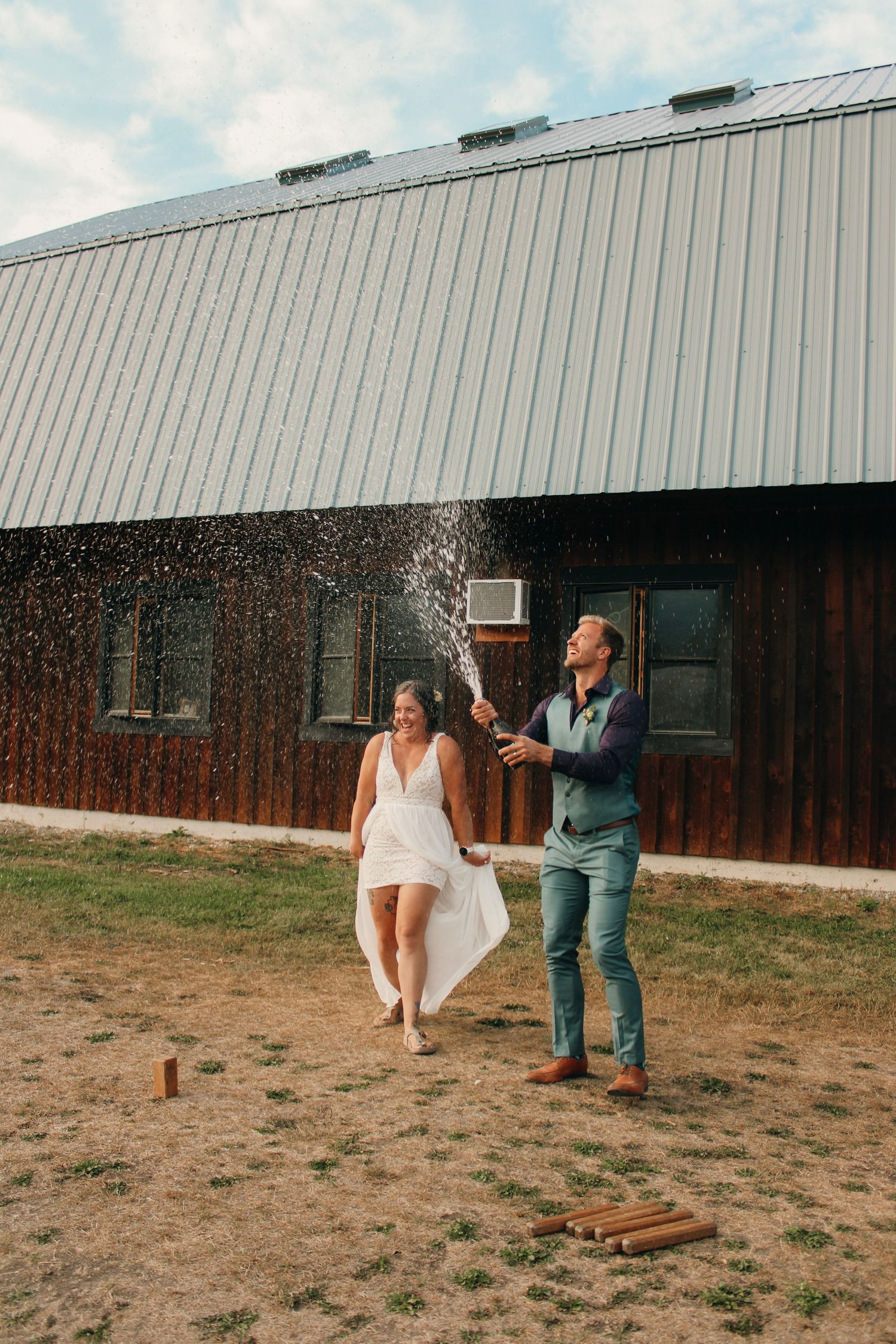 A couple plays a game of cornhole on a grassy field outside a metal building on a sunny day.