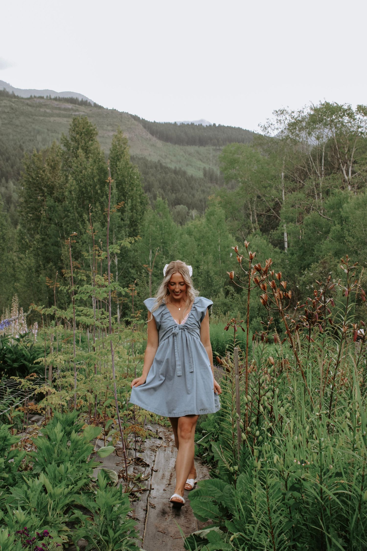 A person in a blue dress explores a wildflower garden with mountains in the background.