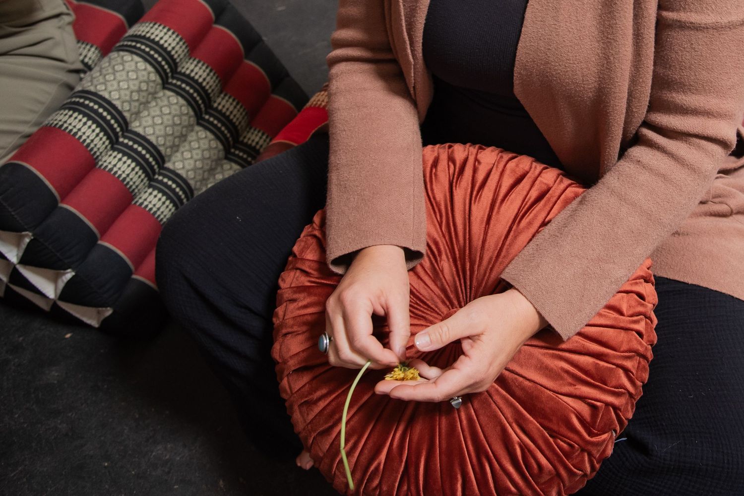 Woman sitting cross-legged holding a round orange velvet pillow, with decorative cushions beside her.
