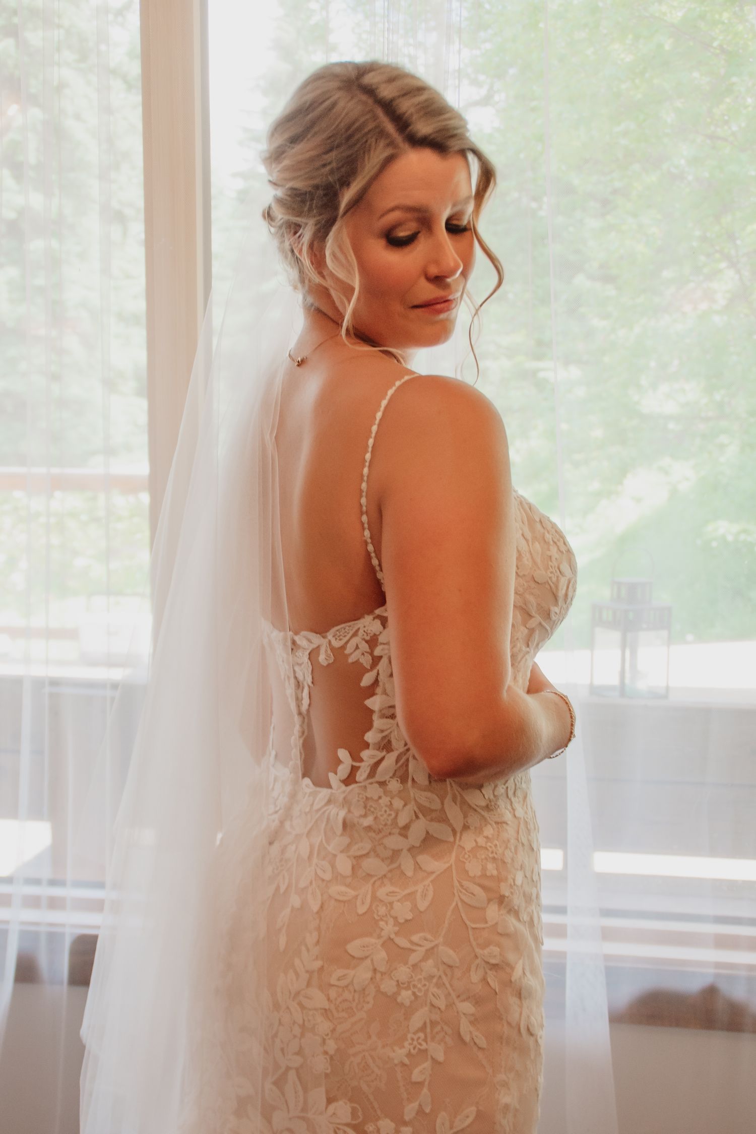 Bride in lace backless wedding gown with veil, posing elegantly by a bright window on her wedding day.