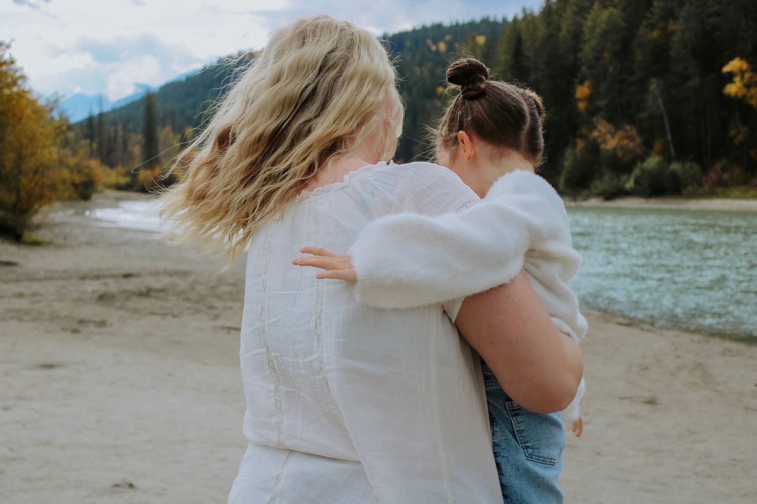 Two women in white sweaters embrace on a scenic riverbank surrounded by mountains and autumn trees.