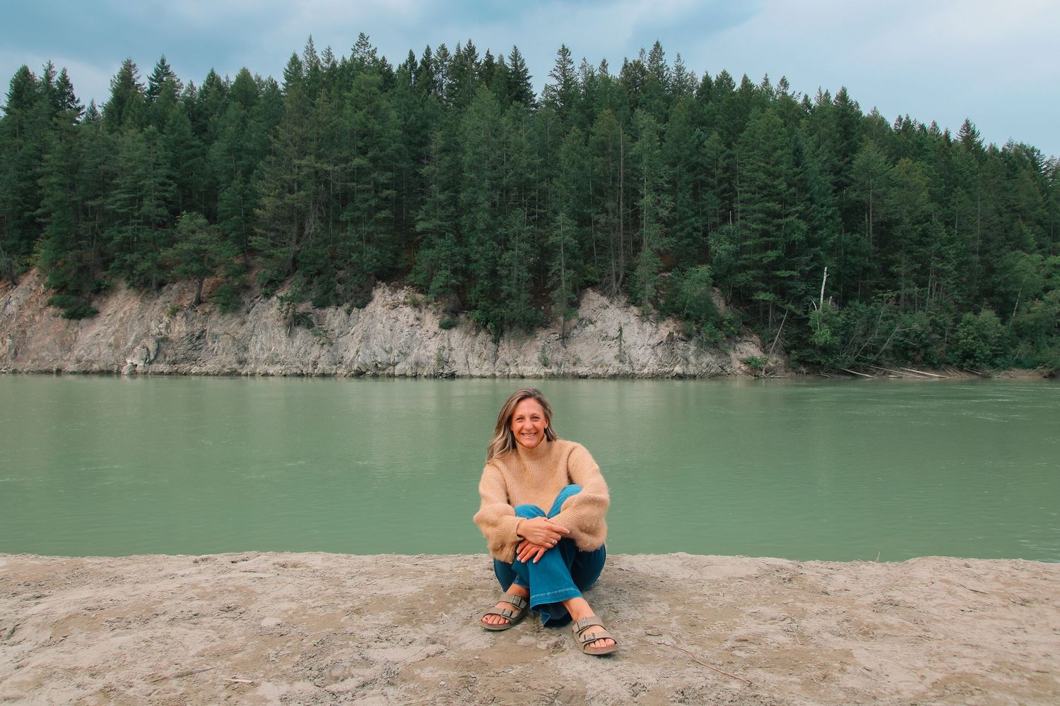 Woman smiling and crouching by a turquoise river with a forested cliff backdrop.