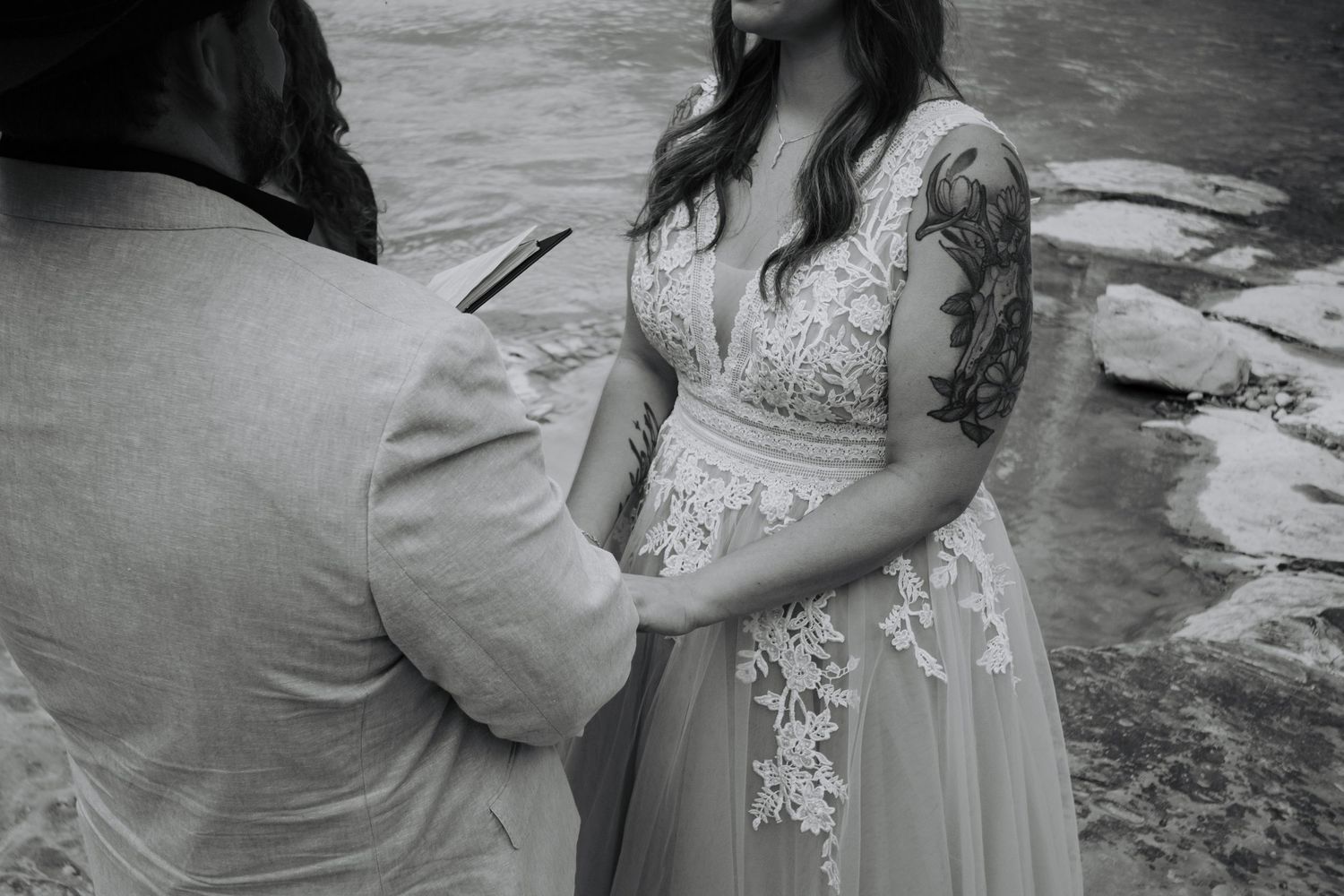 Black and white photo of a bride in lace wedding dress holding hands with officiant during outdoor elopement ceremony.