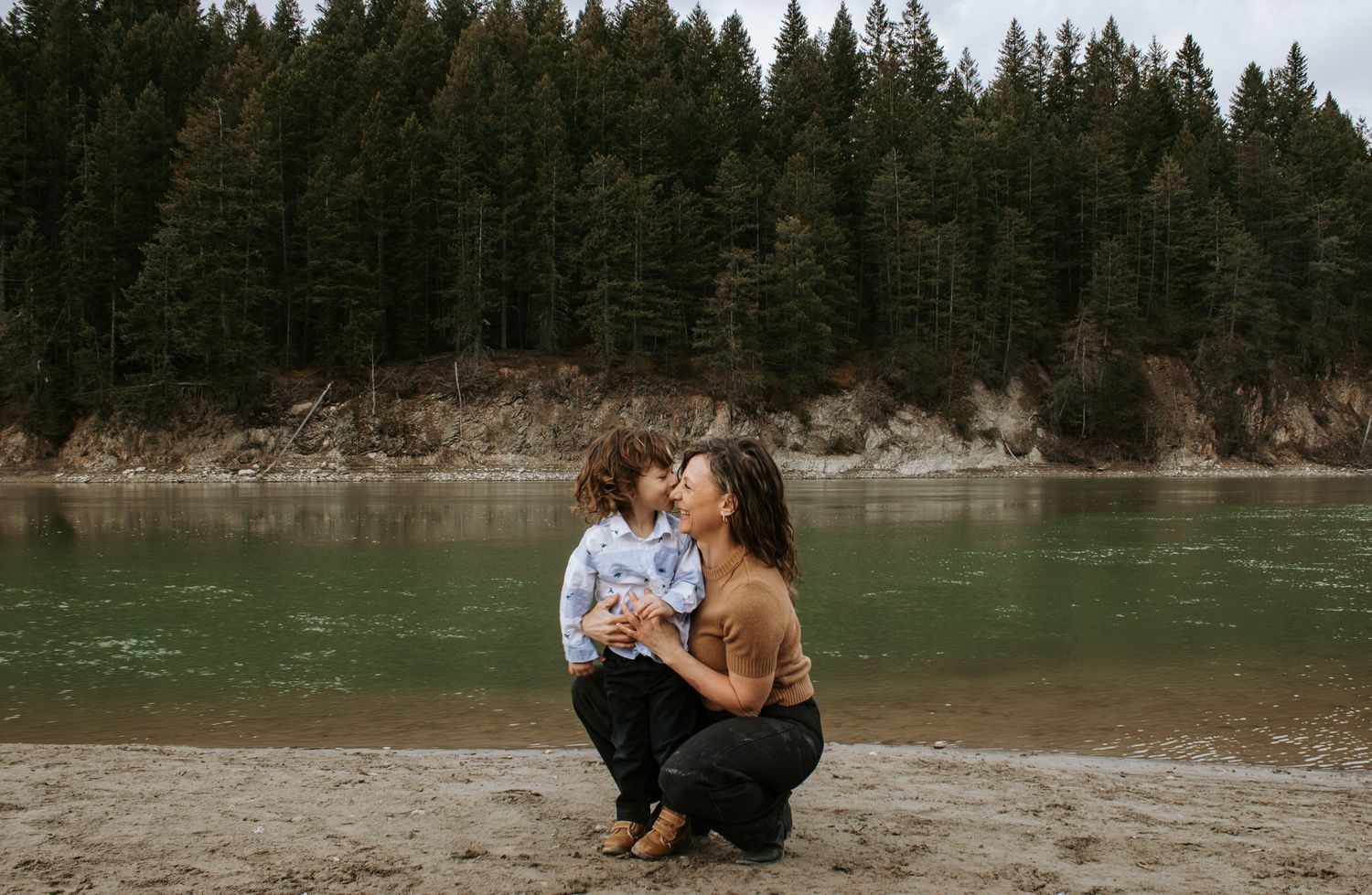 Mother crouching to kiss her toddler on a sandy riverbank with lush evergreen forest in the background.