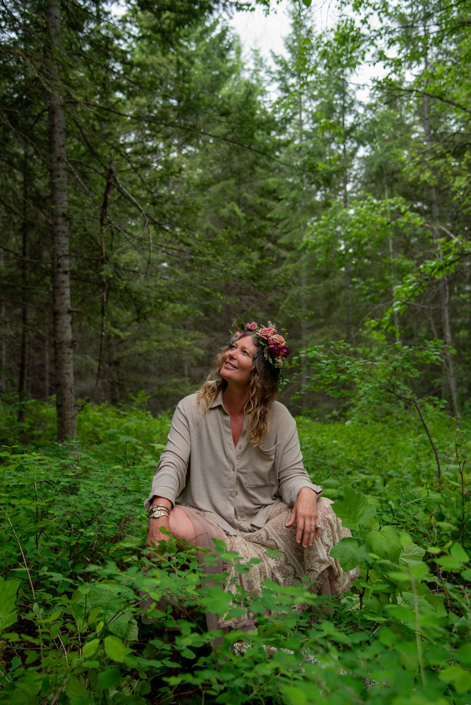 Woman with flower crown sitting peacefully in lush green forest wearing beige dress, surrounded by wild plants and tall pine trees.
