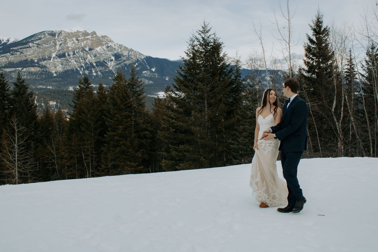 Couple sharing a romantic moment in snowy mountain landscape with evergreen trees and rocky peaks during winter wedding.