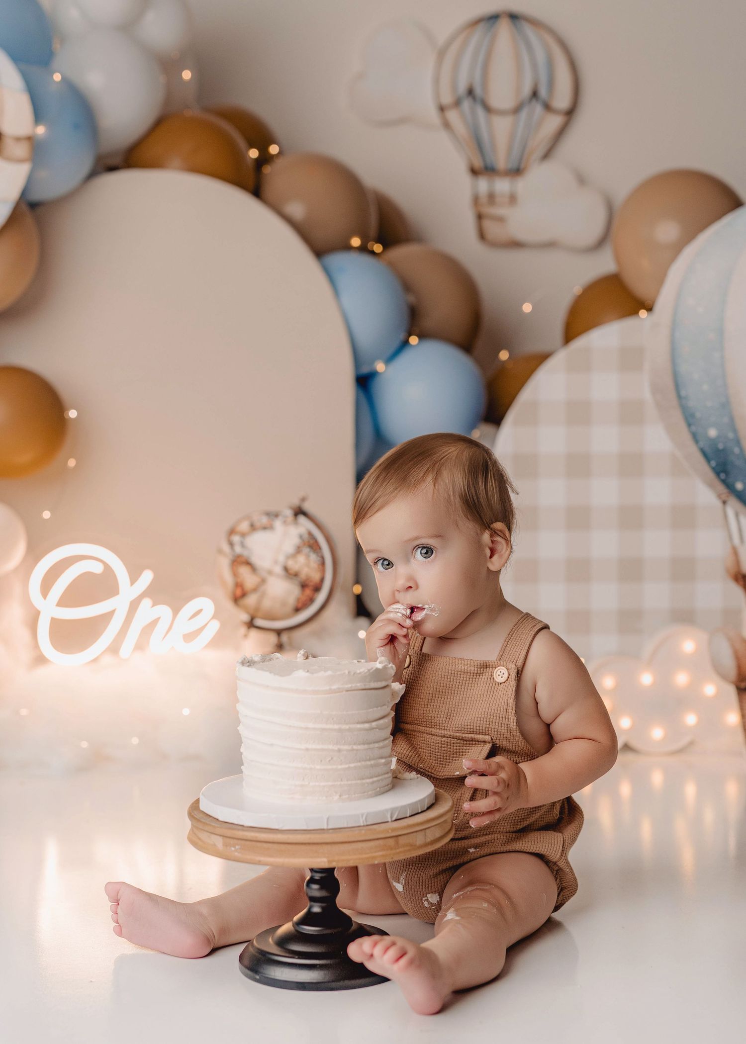 Baby's first birthday cake smash with white cake, blue and tan balloons, hot air balloon backdrop, and One neon sign.