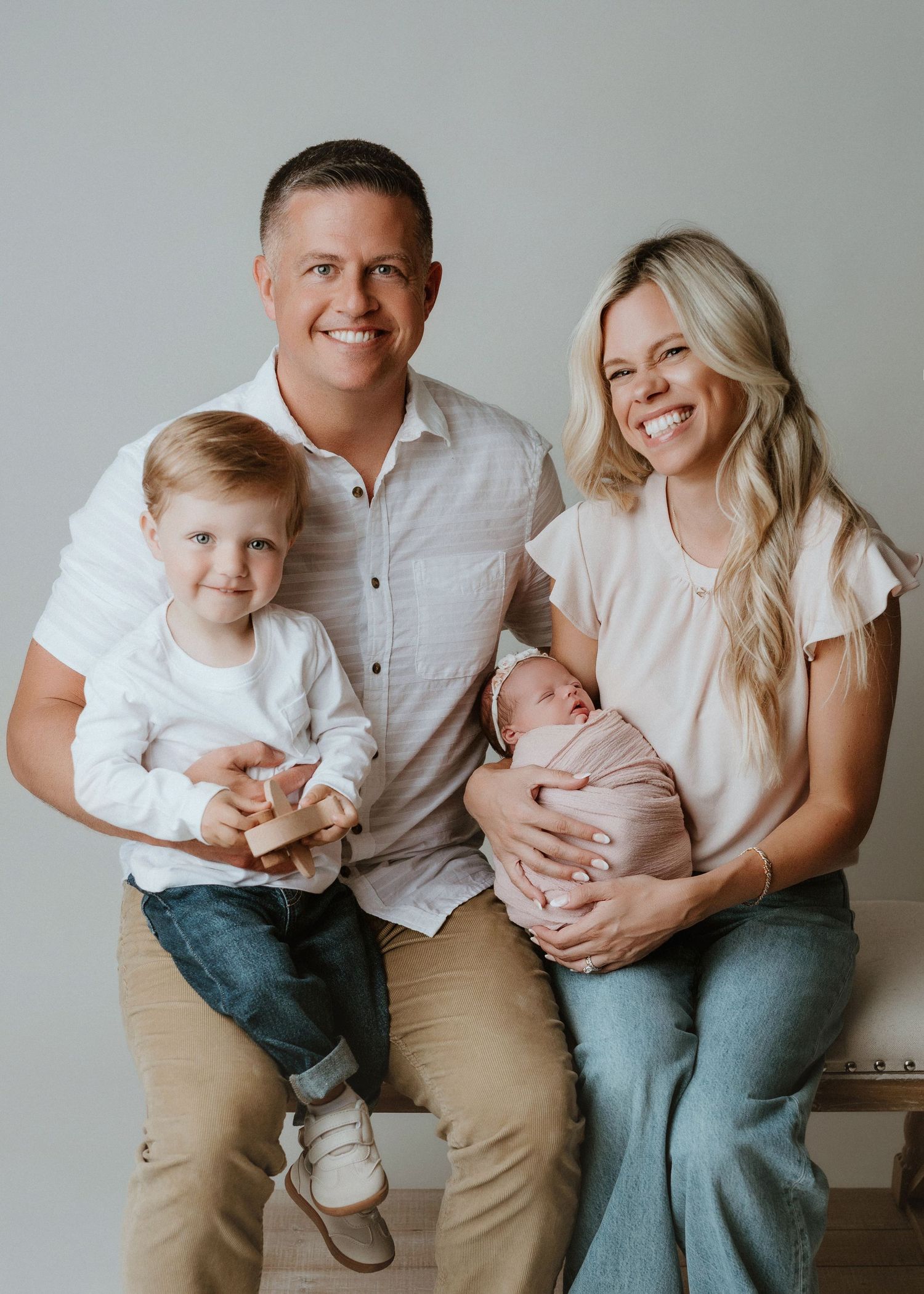 Happy family of four posing for a studio portrait, parents smiling with toddler and newborn baby dressed in soft pink.