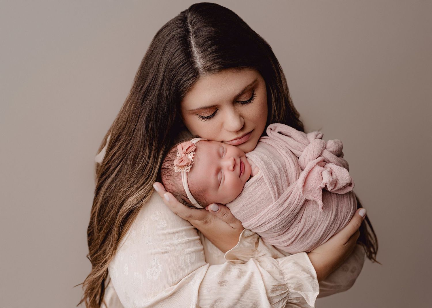 A tender moment between parent and newborn baby wrapped in soft pink swaddle against neutral backdrop.