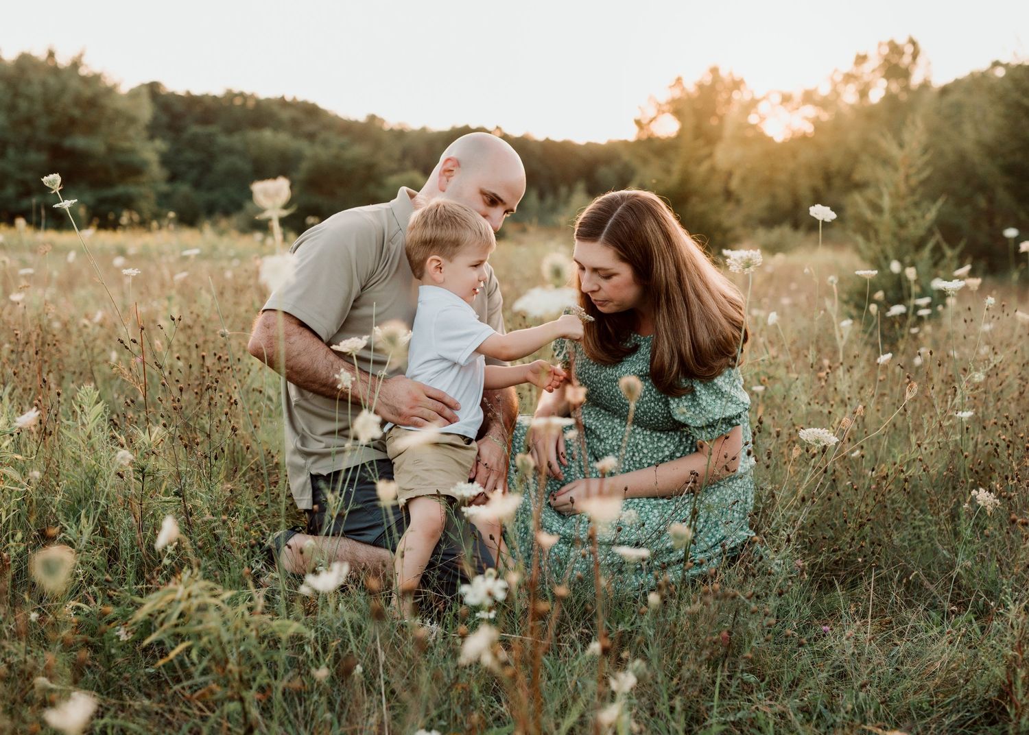 Family with toddler enjoying golden hour in a wildflower meadow, parents kneeling beside child in sunlit field.
