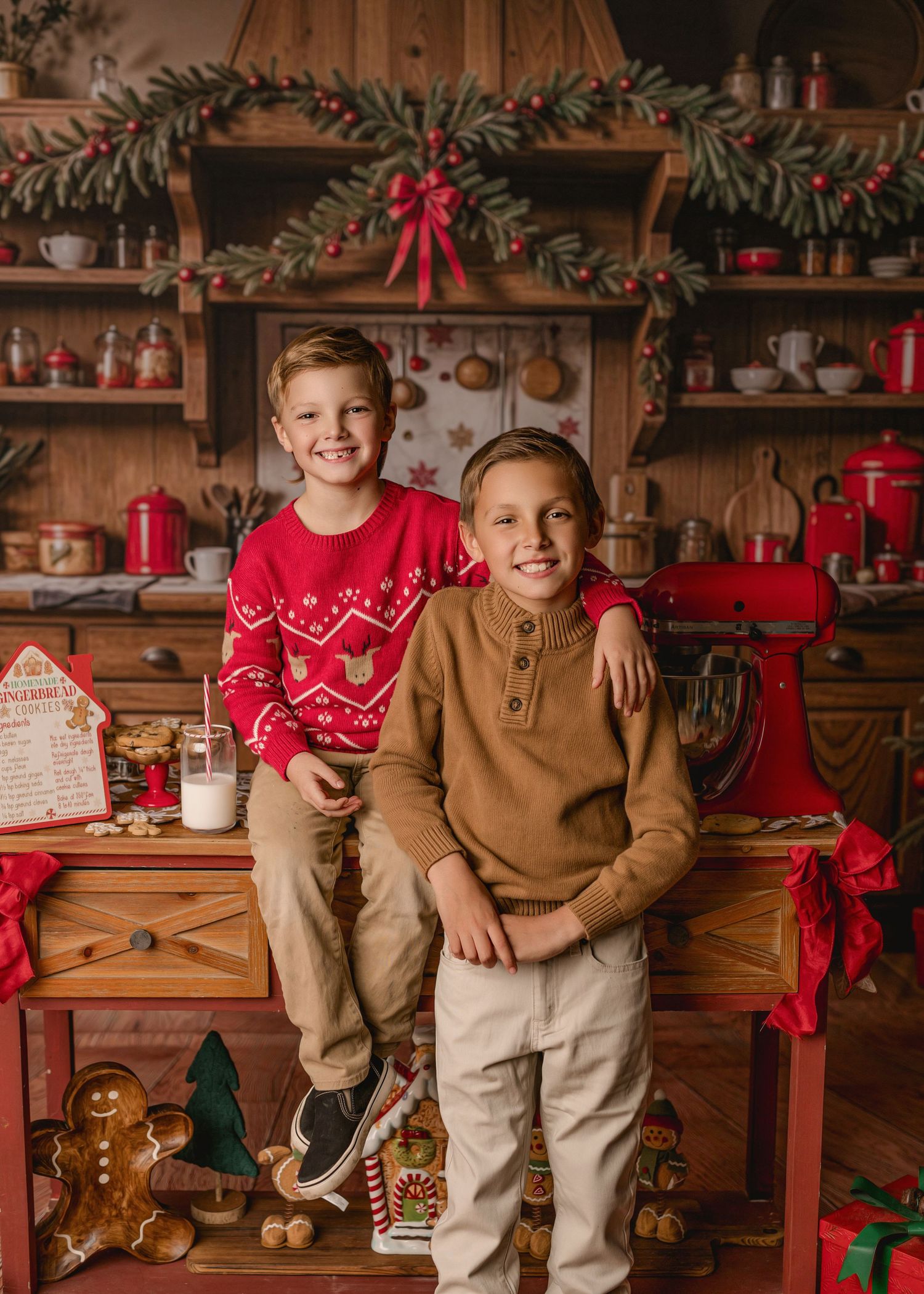 Two children in festive sweaters pose in a rustic Christmas kitchen decorated with garland and holiday decor.