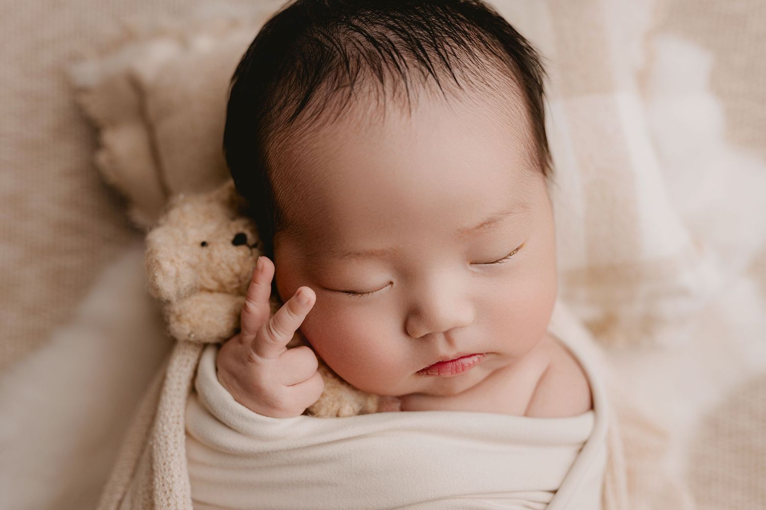 Peaceful newborn baby sleeping swaddled in cream wrap with tiny teddy bear, serene infant photography.