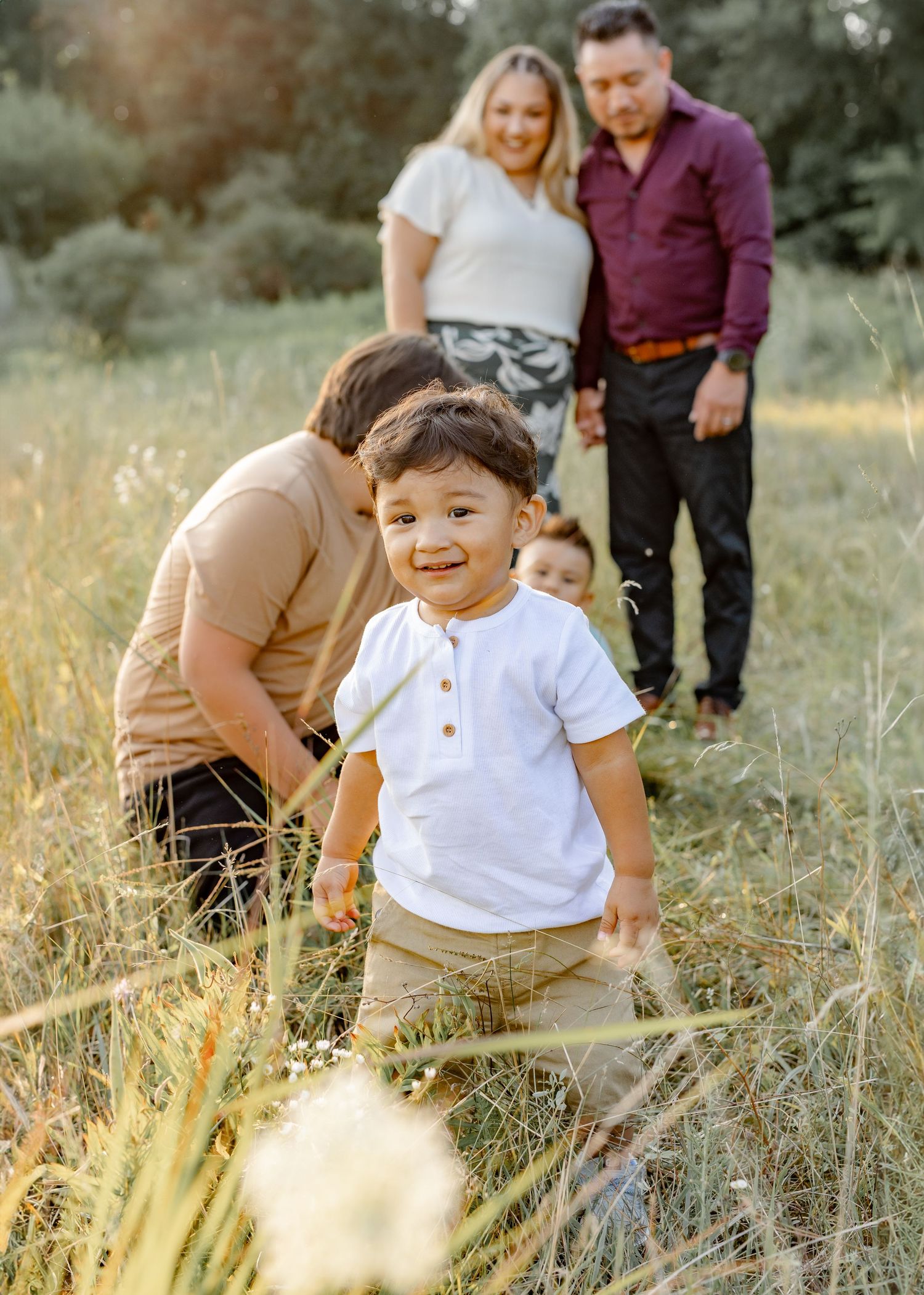 A happy toddler smiles in a sunny field during a family photo session with parents and siblings in the background.