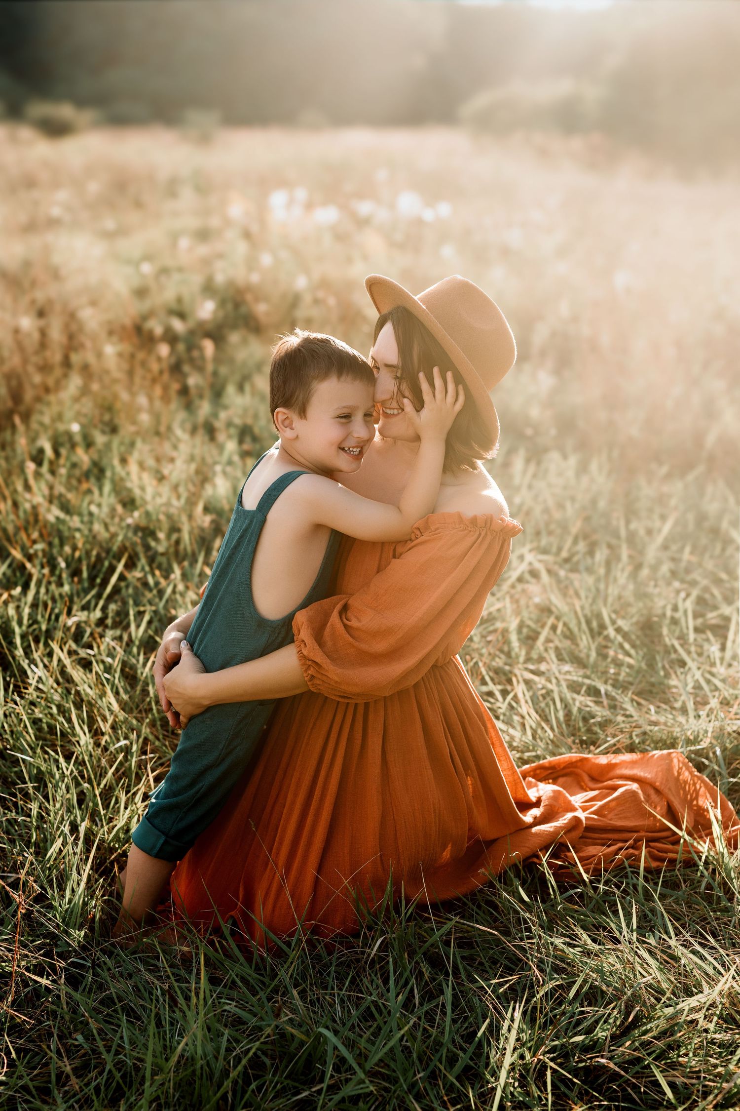 A mother in an orange dress and hat hugs her smiling son in a sunlit golden field.