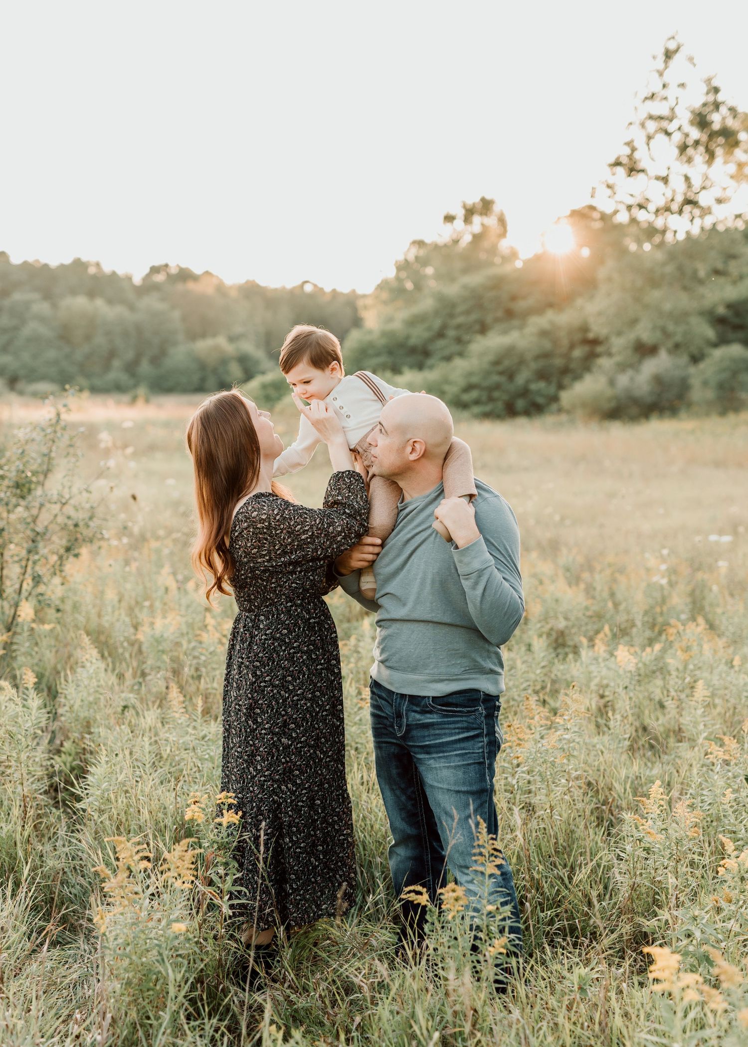 A couple holds their baby aloft in a sunlit meadow, sharing a tender kiss during a golden hour family photo session.