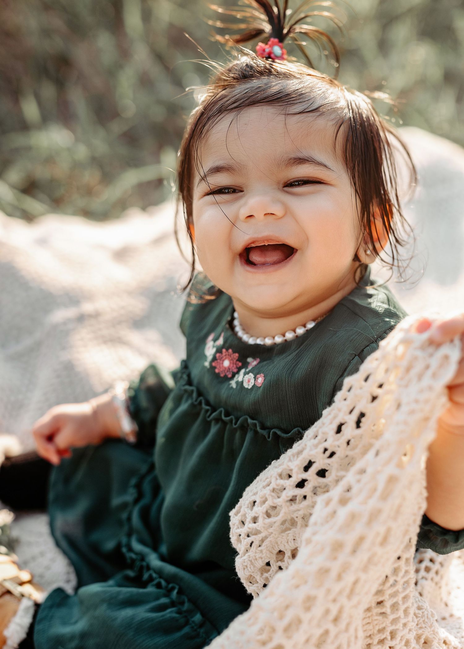 Smiling baby girl in green floral dress with pearl necklace and hair clip, sitting outdoors in natural light.