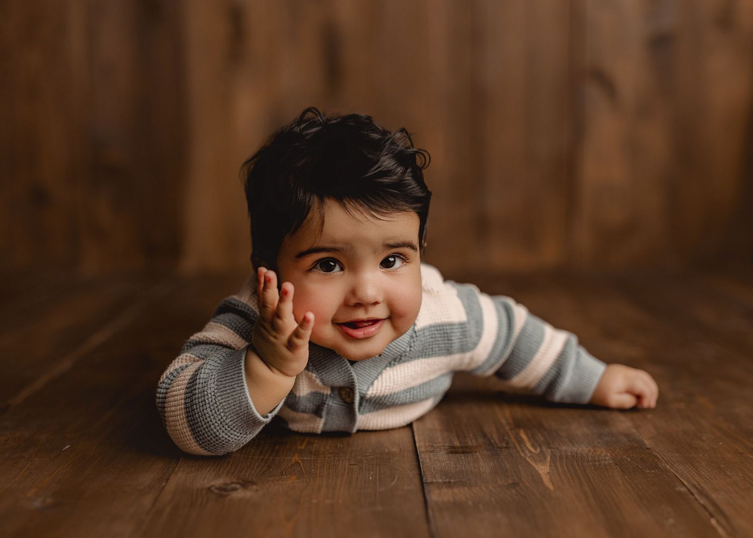 A smiling baby in a striped onesie lies on a wooden floor, reaching one hand forward in a professional studio photo.