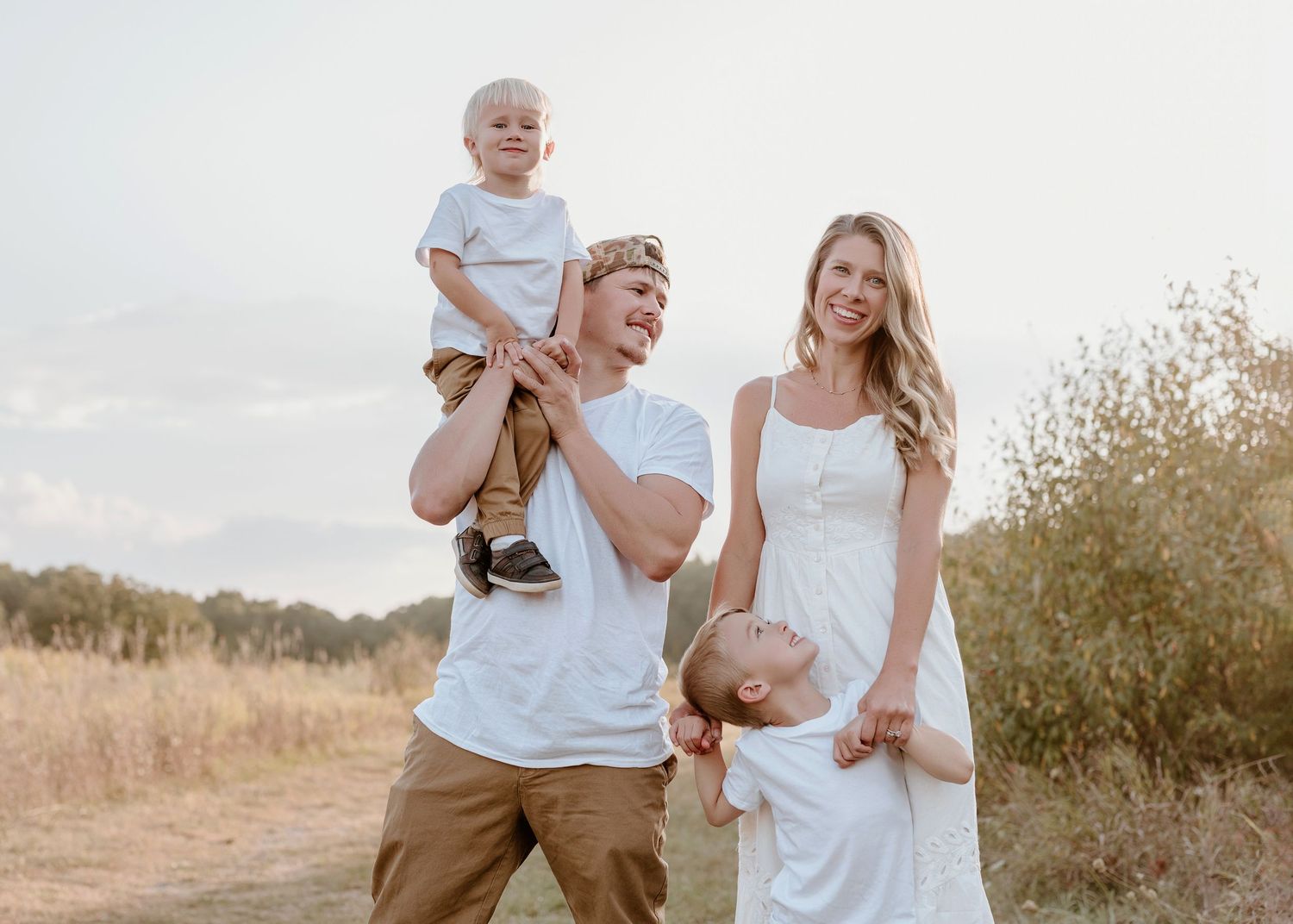 Family of four in white outfits posing outdoors in a sunny field, parents lifting children and smiling Southeast Michigan family photoshoot