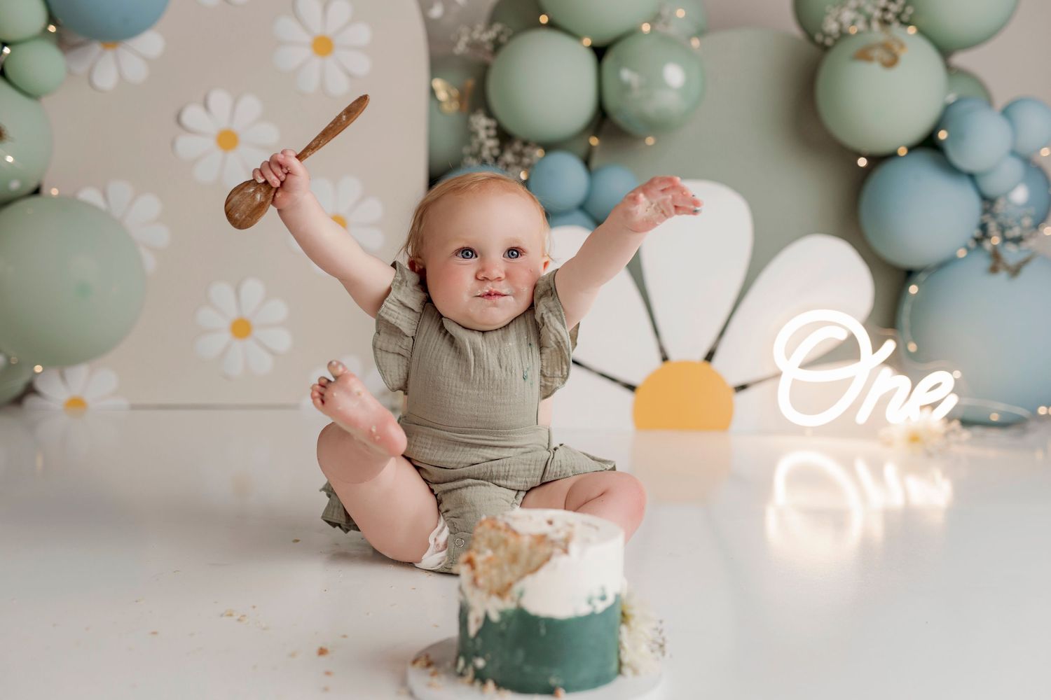 Baby's first birthday cake smash photo with sage green balloons, daisy decorations, and a\u2018One\u2019 neon sign.