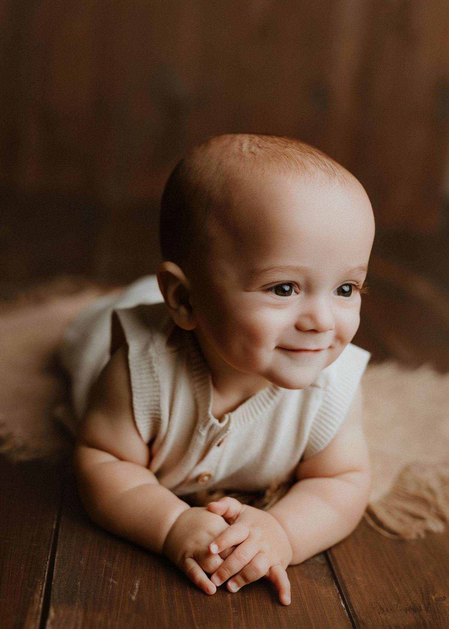 Smiling baby in a cream knit romper lying on a wooden floor with a soft burlap blanket in a warm studio setting.
