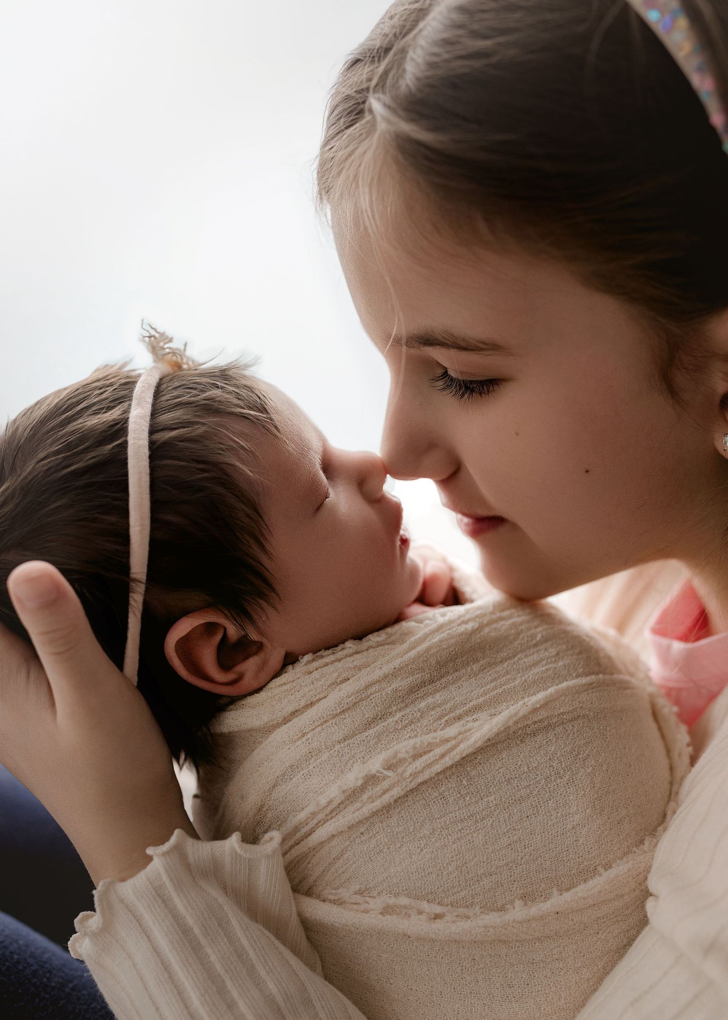 A young girl tenderly holds a swaddled newborn baby close, touching noses in a sweet sibling moment.