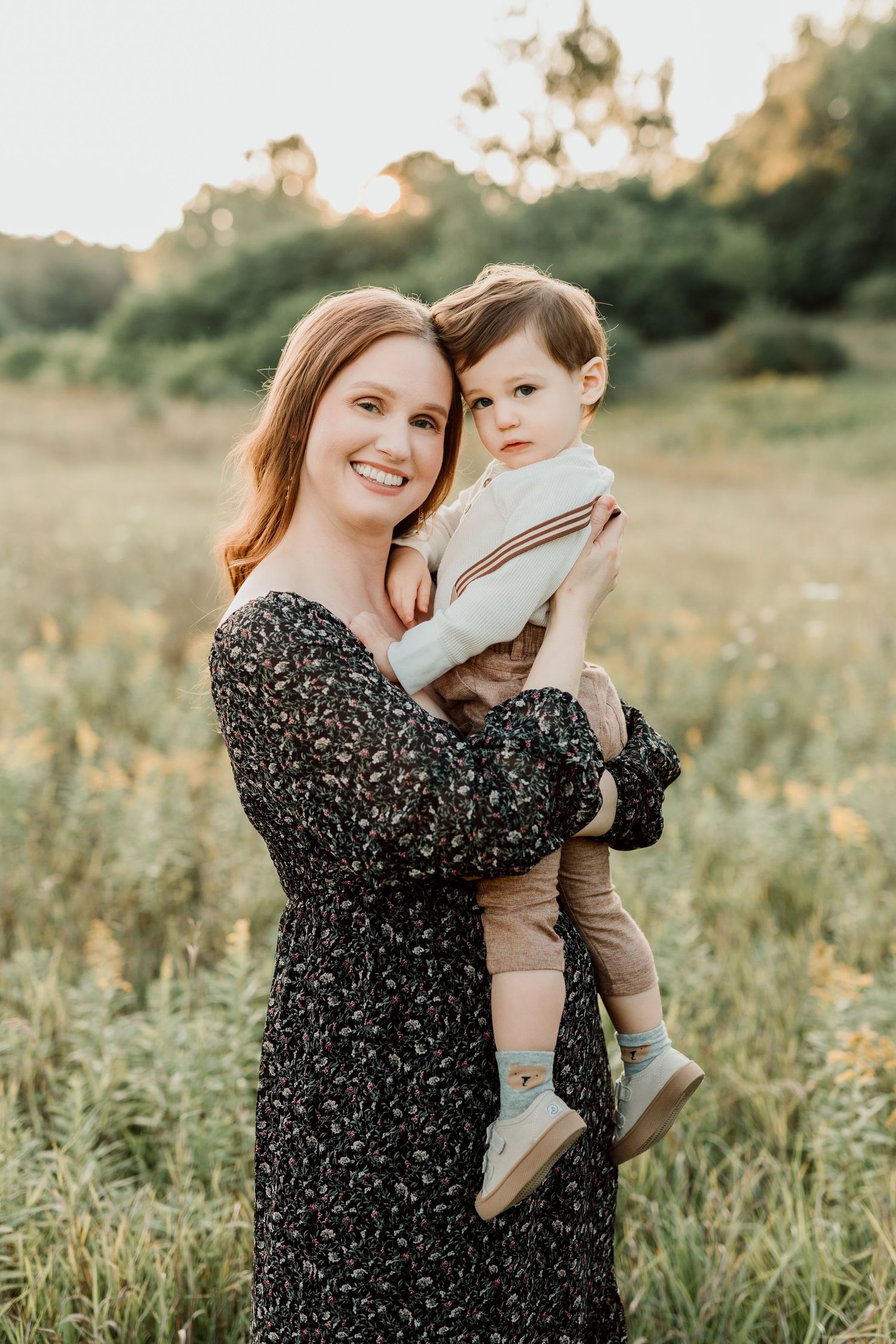 Smiling redhead mother holding toddler in a sunlit meadow, wearing a floral dress during golden hour.