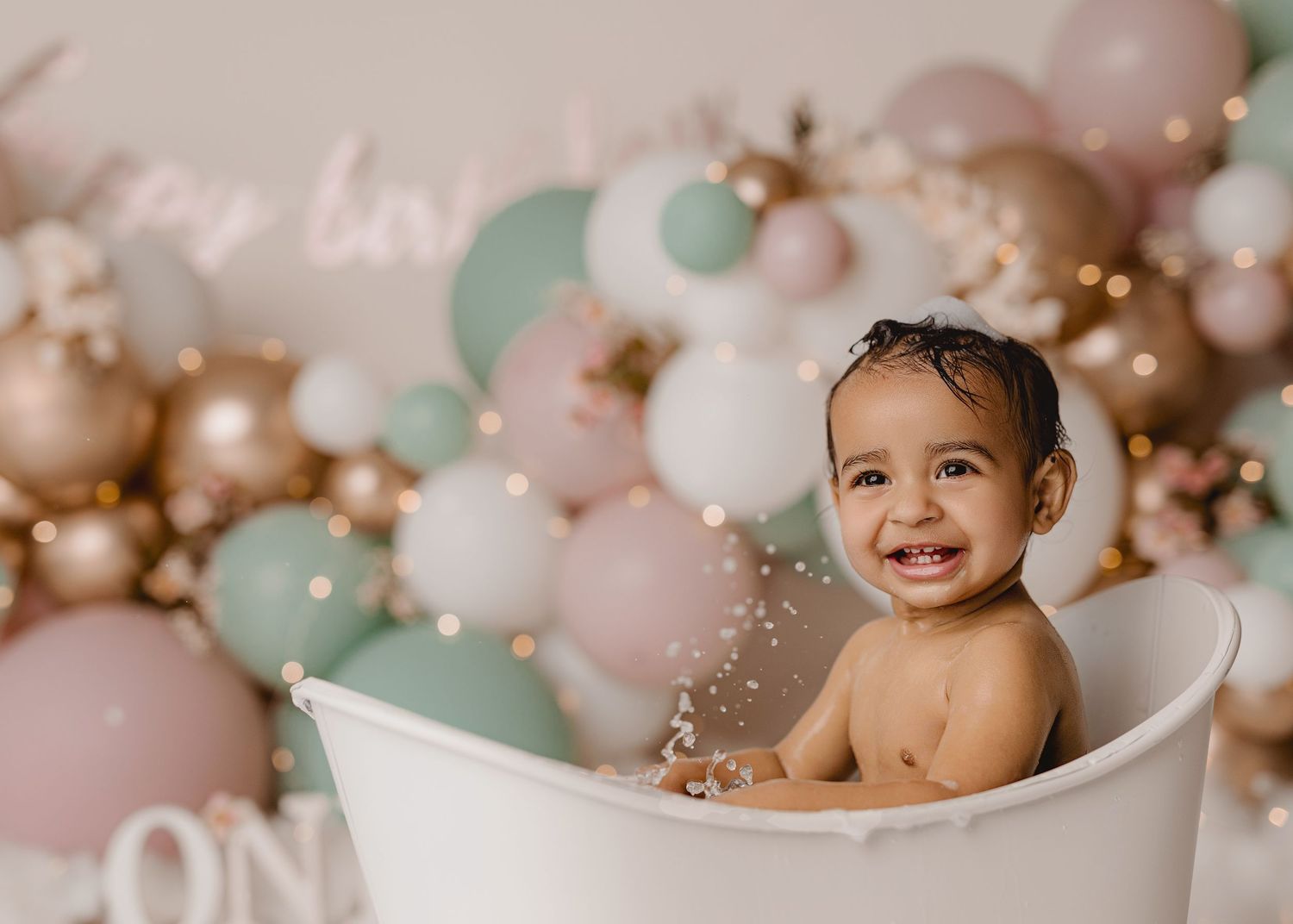 Smiling baby sitting in a white bathtub surrounded by pastel pink, mint, and gold balloons during a first birthday celebration.
