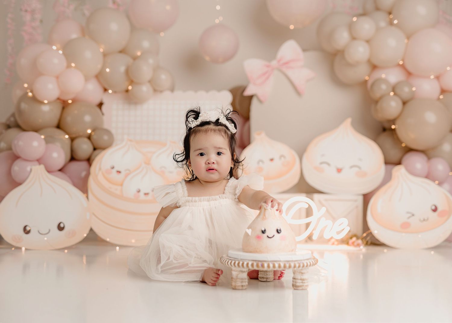 Baby girl in white dress sits before pink balloon arch and dumpling decorations at a first birthday cake smash.